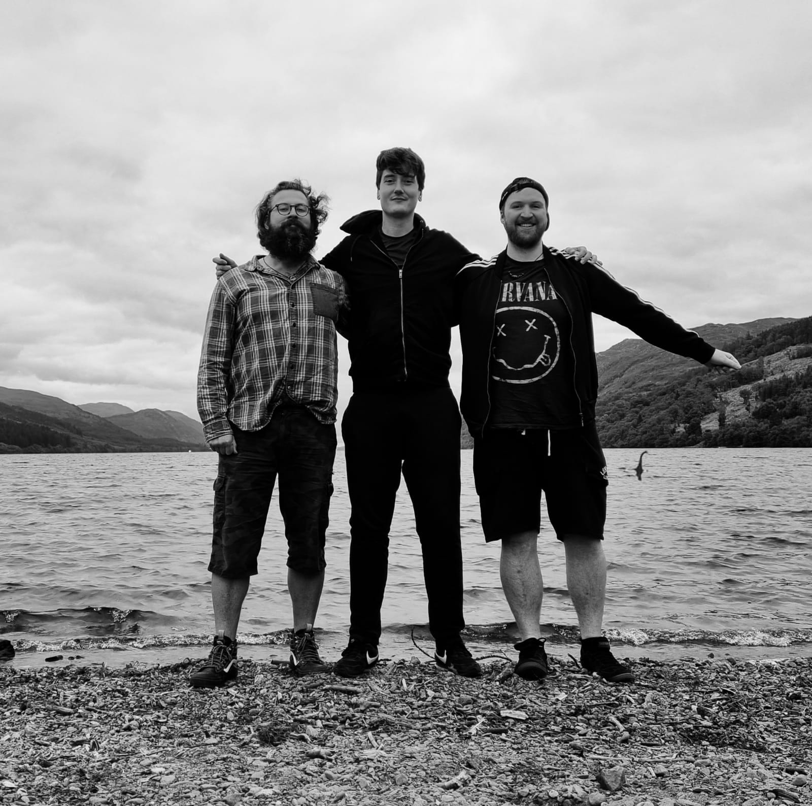 The three band members stand arm in arm on a pebble beach at Loch Ness in black and white, hills behind them and a Nessie-shaped figure in the water