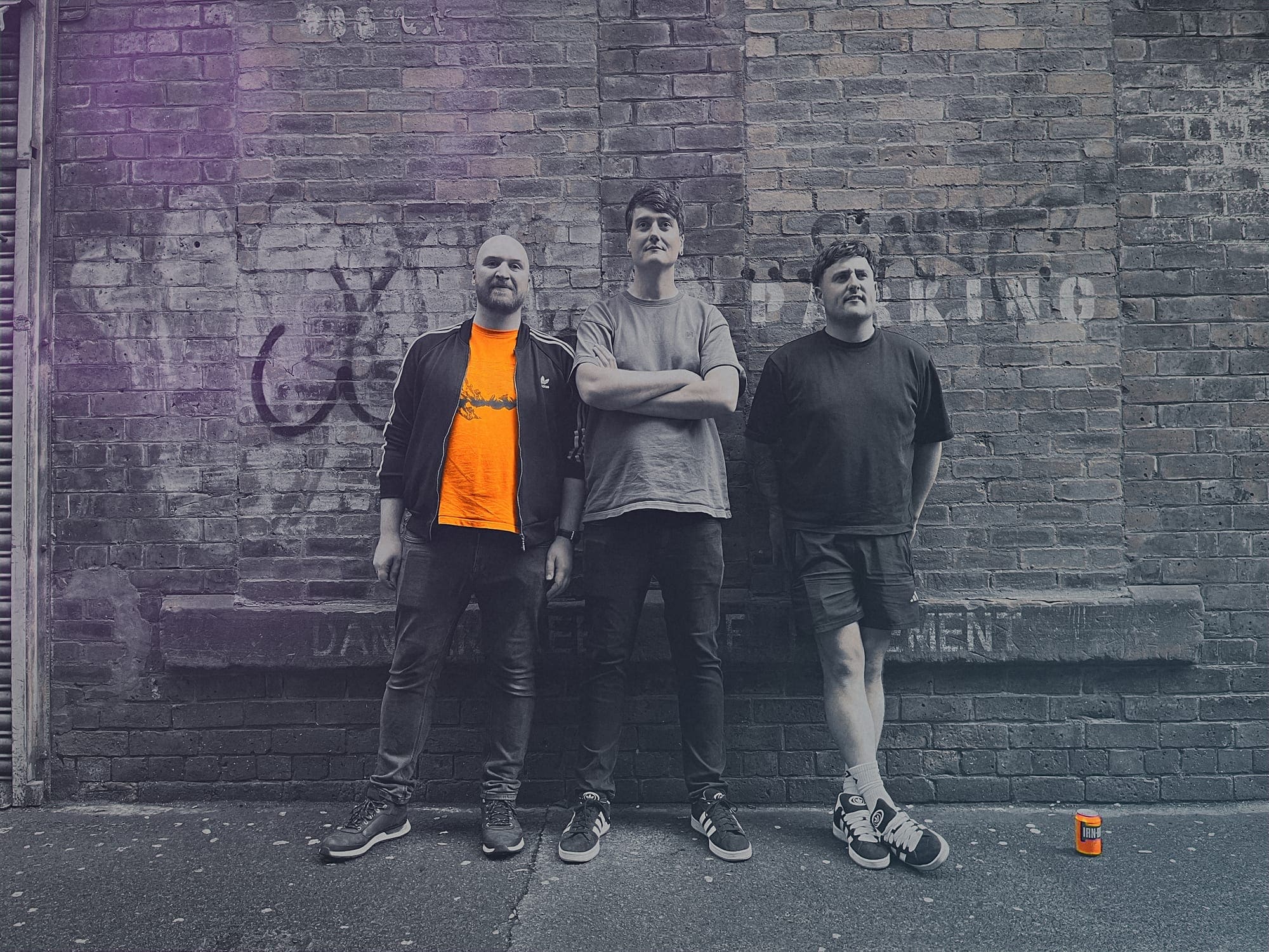 The band stands in front of a graffiti-ed wall in grayscale except for a t-shirt and can of irn-bru